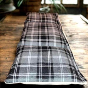 May include: A long, rectangular weighted heating pad featuring a grey, black, and white plaid pattern. The fabric has a soft texture and is stitched into multiple horizontal sections, creating a segmented appearance along the length of the item.