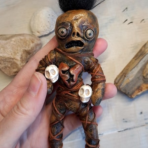 Puede incluir: Muñeca artesanal con un rostro único y expresivo y cabello negro. El cuerpo de la muñeca está envuelto en hilo marrón, con dos pequeñas calaveras en las manos y una abertura en forma de corazón en el pecho. La muñeca mide aproximadamente 15 cm de alto.