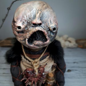 May include: A disturbing doll with a textured face, open mouth, and hollow eyes. It is dressed in black and tan clothing with a fur collar and a pendant. A small glass bottle hangs from the doll's neck, adding to its unsettling appearance.
