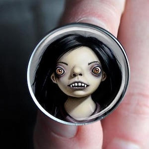 May include: A round, silver-toned ring featuring a miniature portrait of a pale-skinned figure with black hair, red eyes, and sharp teeth. The artwork is set against a dark background, creating a spooky, gothic aesthetic.