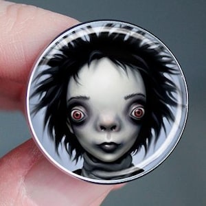 May include: A round, silver-toned button featuring a stylized illustration of a pale-skinned figure with dark, spiky hair and large, red eyes. The button is held between fingers against a blurred background.