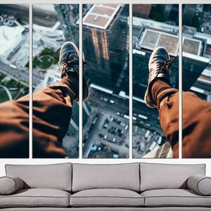 Puede incluir: Las piernas de una persona cuelgan sobre el borde de un edificio, con el horizonte de la ciudad visible debajo. La persona lleva pantalones marrones y zapatillas negras.