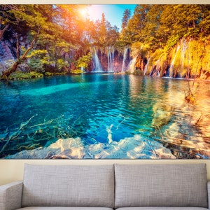 May include: A scenic landscape photograph featuring a turquoise blue lake with waterfalls cascading down a rocky cliffside. Lush green trees and foliage surround the lake, creating a tranquil and picturesque scene.
