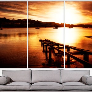 May include: A sunset over a body of water with a wooden pier extending into the water. The sky is a vibrant orange and yellow, and the water is a calm, reflective orange.