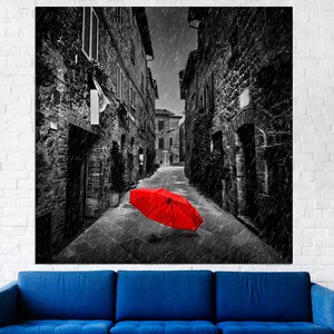 May include: A black and white photograph of a narrow street in a European city. The street is empty except for a single red umbrella lying on the ground. The photo is taken from a low angle, looking up the street.