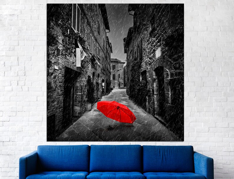 May include: A black and white photograph of a narrow street in a European city. The street is empty except for a single red umbrella lying on the ground. The photo is taken from a low angle, looking up the street.