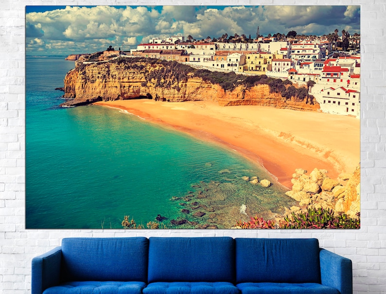 May include: A scenic view of a white-washed village perched on a cliff overlooking a turquoise bay with a sandy beach. The sky is a mix of blue and white clouds.