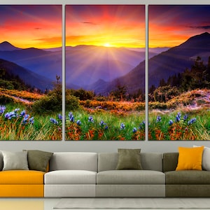 May include: A three-panel canvas print depicting a vibrant sunset over a mountain range. The sky is a mix of orange, pink, and purple, with rays of light emanating from the sun. The mountains are a deep blue, and the foreground is a field of green grass with purple wildflowers.