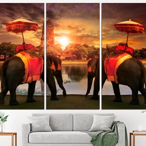 May include: Three elephants adorned with red and gold fabric walk along a lake with a temple in the background. The sky is a vibrant orange and yellow sunset.