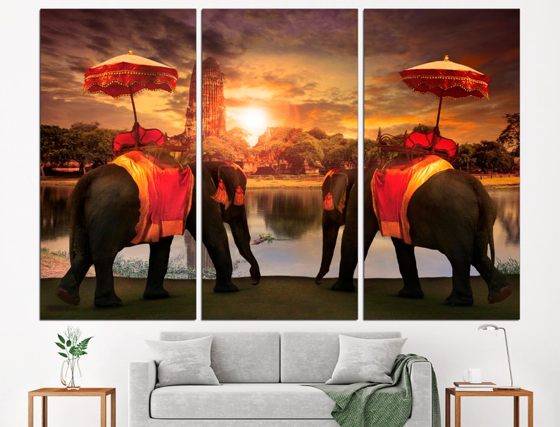 May include: Three elephants adorned with red and gold fabric walk along a lake with a temple in the background. The sky is a vibrant orange and yellow sunset.