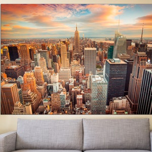 May include: A panoramic view of the New York City skyline at sunset. The Empire State Building is visible in the distance, with other skyscrapers and buildings in the foreground. The sky is a vibrant orange and pink, with clouds casting long shadows over the city.