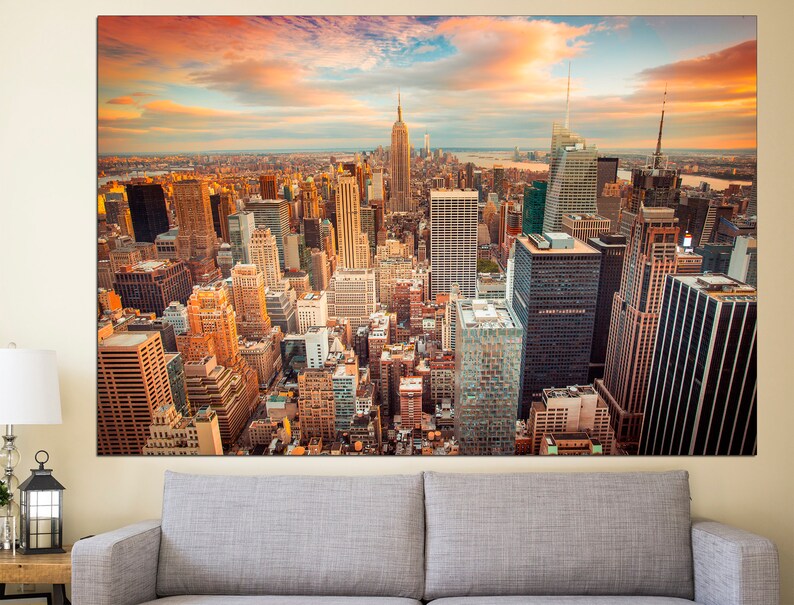 May include: A panoramic view of the New York City skyline at sunset. The Empire State Building is visible in the distance, with other skyscrapers and buildings in the foreground. The sky is a vibrant orange and pink, with clouds casting long shadows over the city.