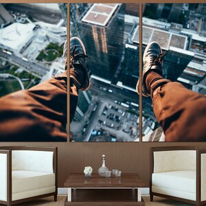 Puede incluir: Las piernas de una persona cuelgan sobre el borde de un edificio, con los pies en zapatillas negras. La ciudad de abajo es visible, con edificios y calles.