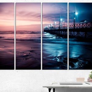 May include: A four-panel canvas print of a beach scene at dusk. The sky transitions from pink to blue, reflecting in the water and wet sand. A wooden pier with lights extends into the ocean. The overall mood is serene and atmospheric.