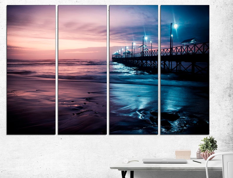 May include: A four-panel canvas print of a beach scene at dusk. The sky transitions from pink to blue, reflecting in the water and wet sand. A wooden pier with lights extends into the ocean. The overall mood is serene and atmospheric.