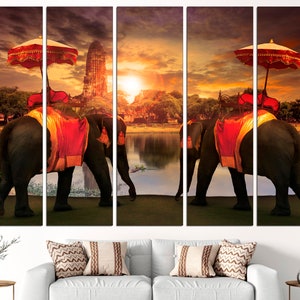 May include: A multi-panel canvas print featuring two elephants adorned with red and gold fabric, walking towards a temple in the distance. The elephants are carrying ornate canopies with red and white umbrellas. The scene is set at sunset, with a golden sky and a reflecting lake.