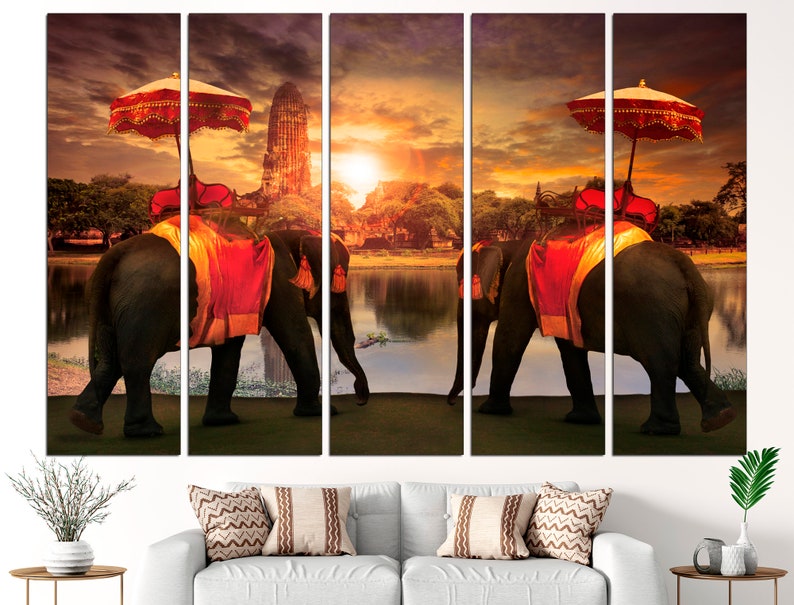 May include: A multi-panel canvas print featuring two elephants adorned with red and gold fabric, walking towards a temple in the distance. The elephants are carrying ornate canopies with red and white umbrellas. The scene is set at sunset, with a golden sky and a reflecting lake.