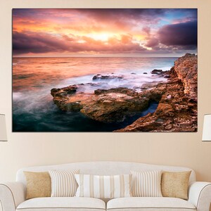 Pode incluir: Uma fotografia panor&acirc;mica de uma costa rochosa com um c&eacute;u de p&ocirc;r do sol espetacular. O c&eacute;u &eacute; uma mistura de tons rosados, laranja e roxos, com nuvens que projetam longas sombras sobre a &aacute;gua. As ondas quebram contra as rochas, criando uma sensa&ccedil;&atilde;o de movimento e energia.