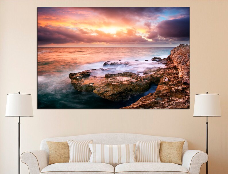 Pode incluir: Uma fotografia panor&acirc;mica de uma costa rochosa com um c&eacute;u de p&ocirc;r do sol espetacular. O c&eacute;u &eacute; uma mistura de tons rosados, laranja e roxos, com nuvens que projetam longas sombras sobre a &aacute;gua. As ondas quebram contra as rochas, criando uma sensa&ccedil;&atilde;o de movimento e energia.