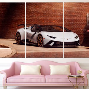 May include: A white Lamborghini sports car parked in front of a brick wall. The car has a black interior and black wheels with gold accents.