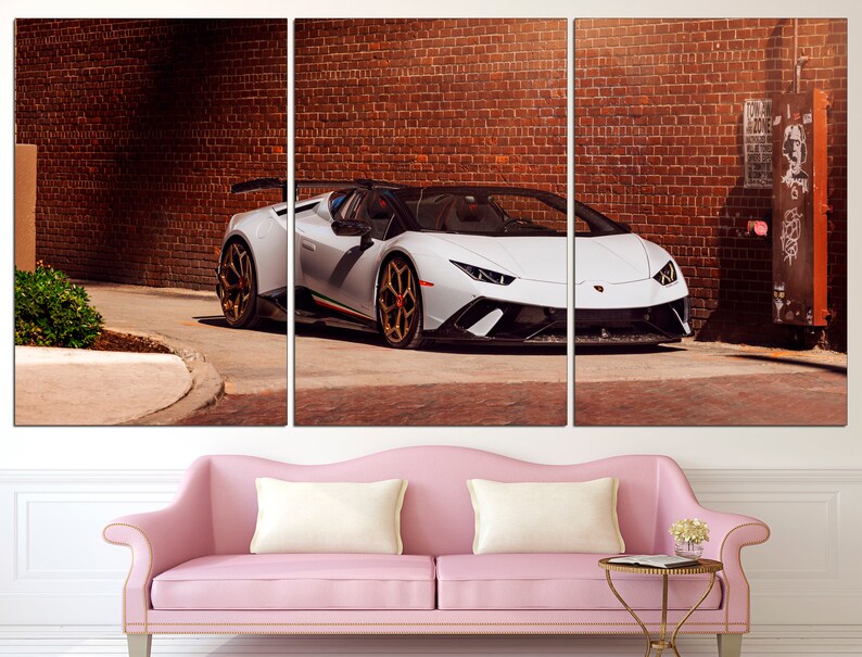 May include: A white Lamborghini sports car parked in front of a brick wall. The car has a black interior and black wheels with gold accents.