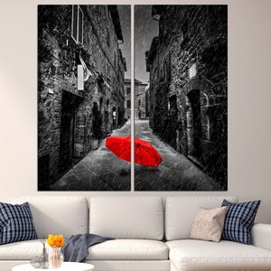 May include: A black and white photograph of a narrow street in a European city. The street is empty except for a bright red umbrella lying on the ground in the middle of the street.