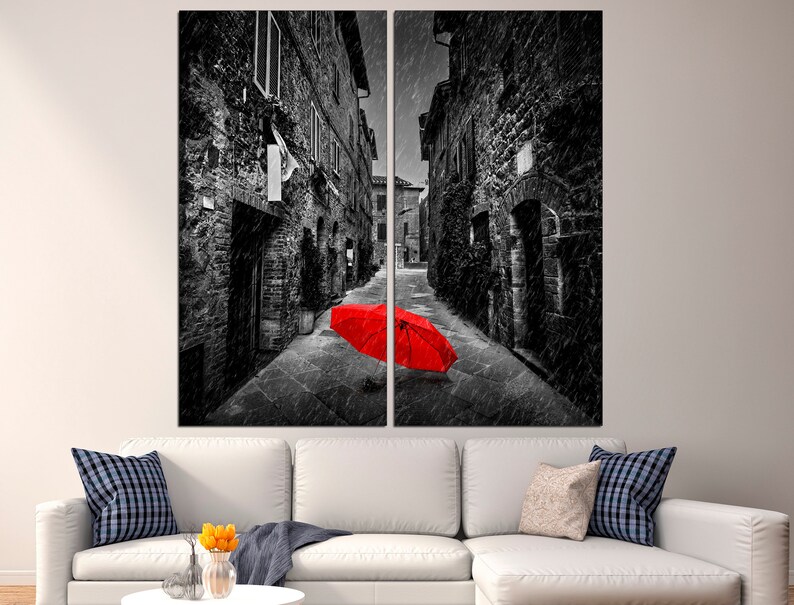 May include: A black and white photograph of a narrow street in a European city. The street is empty except for a bright red umbrella lying on the ground in the middle of the street.