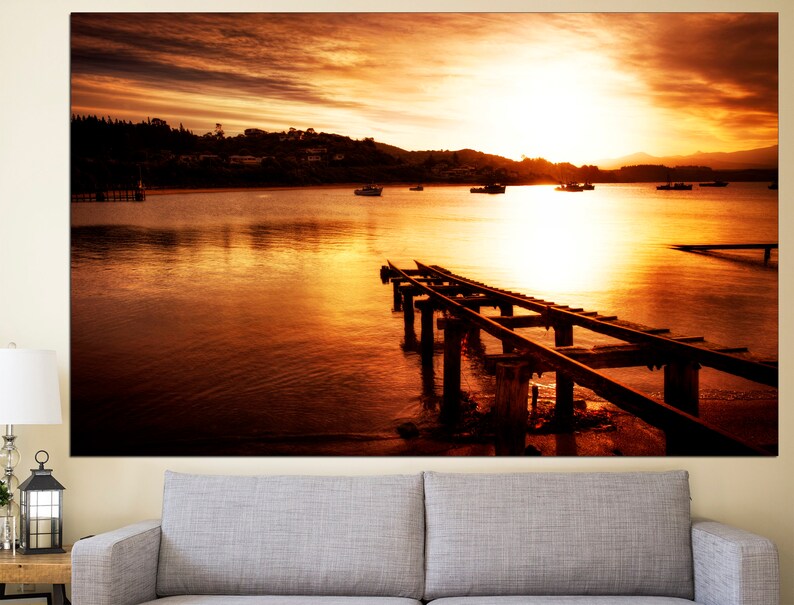 May include: A wooden pier extends into a calm body of water at sunset. The sky is a vibrant orange and yellow, with the sun setting in the distance. The water reflects the colors of the sky, creating a beautiful and serene scene.