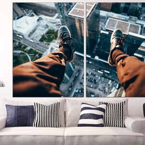 Puede incluir: Una imagen dividida en dos partes de las piernas de una persona colgando del borde de un edificio alto. La persona lleva pantalones marrones y zapatillas negras. El horizonte de la ciudad es visible en el fondo.