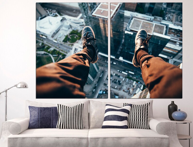 Puede incluir: Una imagen dividida en dos partes de las piernas de una persona colgando del borde de un edificio alto. La persona lleva pantalones marrones y zapatillas negras. El horizonte de la ciudad es visible en el fondo.