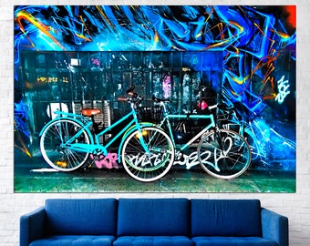 May include: Two teal blue bicycles parked in front of a wall with colourful graffiti. The graffiti features blue, orange, and yellow paint.