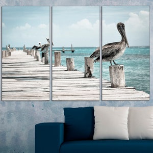 May include: A triptych of three canvas prints featuring a pelican perched on a wooden post over a turquoise ocean. The pelican is looking to the right, and the wooden pier extends into the distance.