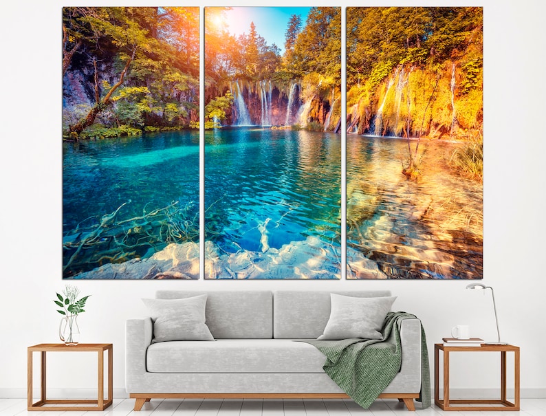 May include: A three-panel canvas print featuring a scenic landscape with a waterfall cascading into a turquoise lake surrounded by lush green trees. The water is clear and reflects the sunlight. The trees are in shades of green and yellow, creating a vibrant autumnal scene.