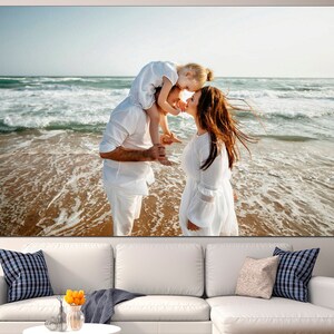 May include: A family of three on a beach, the parents are holding their young daughter, all three are wearing white clothing and smiling at each other.