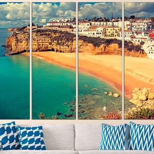 May include: A panoramic view of a coastal town in Portugal. The image shows a white-washed town built on a cliff overlooking a sandy beach and turquoise water. The sky is blue with white clouds.