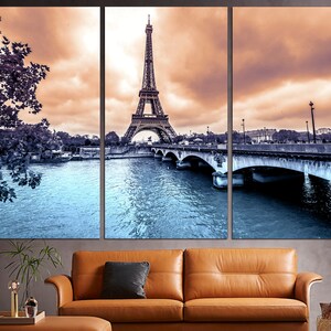May include: A panoramic view of the Eiffel Tower in Paris, France. The tower is in the center of the image, with a bridge and river in the foreground. The sky is a soft orange and pink, with clouds.