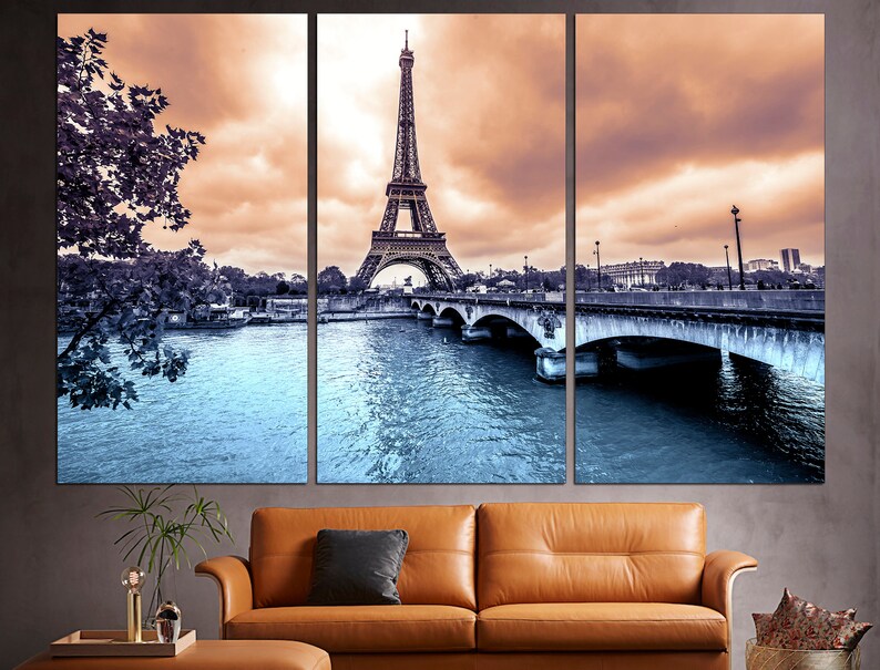 May include: A panoramic view of the Eiffel Tower in Paris, France. The tower is in the center of the image, with a bridge and river in the foreground. The sky is a soft orange and pink, with clouds.