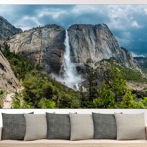 May include: A panoramic view of a waterfall cascading down a rocky cliff face in Yosemite National Park. Lush green trees and a cloudy sky complete the scene.