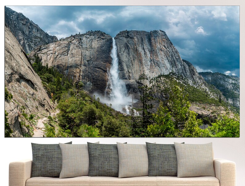 May include: A panoramic view of a waterfall cascading down a rocky cliff face in Yosemite National Park. Lush green trees and a cloudy sky complete the scene.