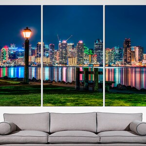 Puede incluir: Una ciudad nocturna con una vista del horizonte de San Diego reflejada en el agua. Una sola farola se encuentra en primer plano, proyectando un c&aacute;lido resplandor sobre la hierba verde.