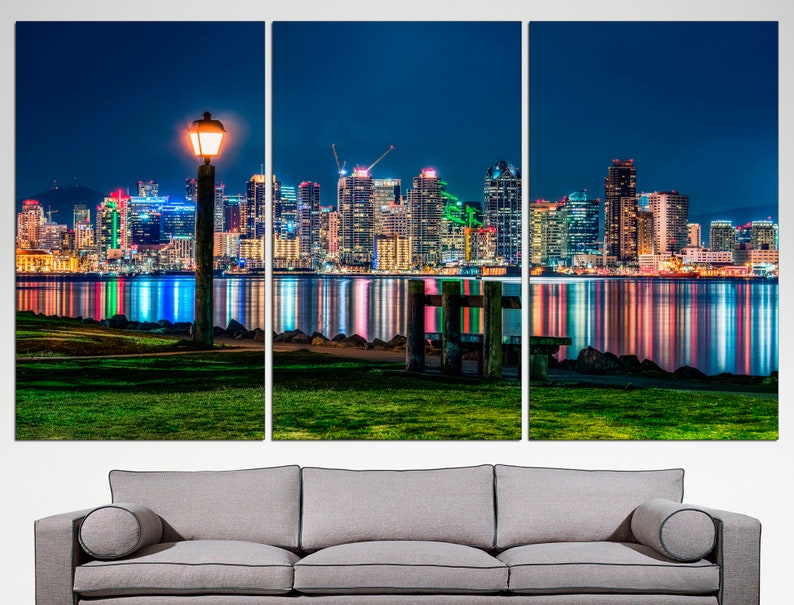 Puede incluir: Una ciudad nocturna con una vista del horizonte de San Diego reflejada en el agua. Una sola farola se encuentra en primer plano, proyectando un c&aacute;lido resplandor sobre la hierba verde.
