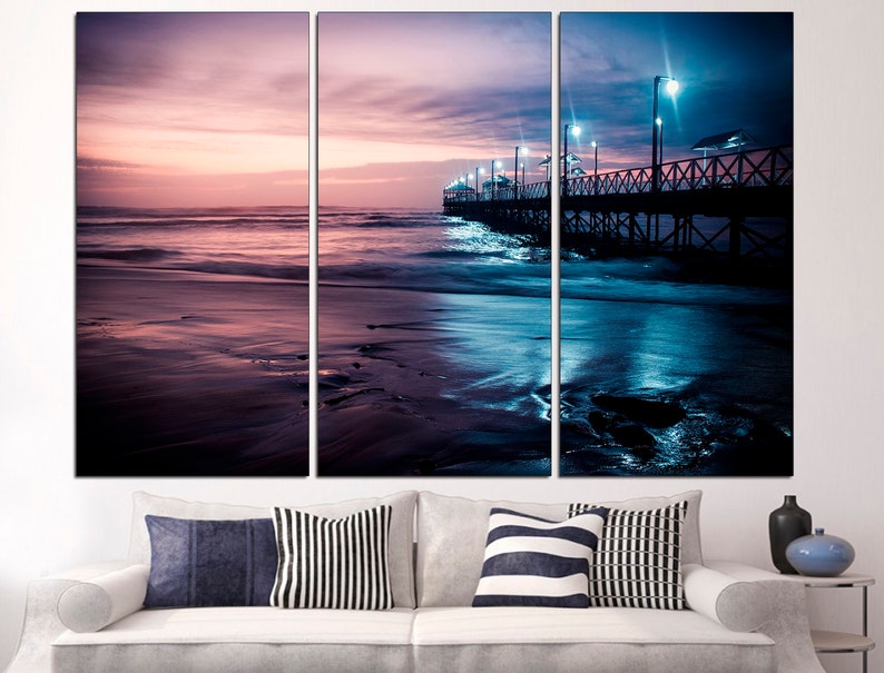 May include: A triptych canvas print depicting a serene beach scene at dusk. The artwork features a pier extending into the ocean under a twilight sky, with shades of purple, pink, and blue. The image is mounted above a white sofa with decorative pillows.