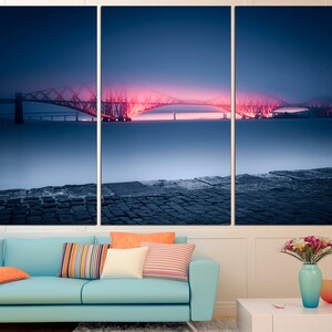 May include: A three-panel canvas print featuring a bridge lit up in pink and red against a dark blue sky. The bridge is reflected in the water below, which is covered in a light fog. The bottom of the image shows a stone walkway.