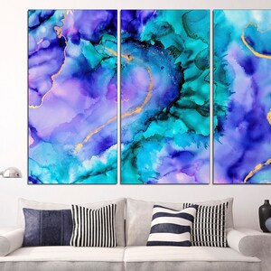 May include: Abstract wall art in a triptych arrangement. The artwork features a blend of blue, teal, and purple hues with gold accents. The fluid design creates a watercolor effect, adding a touch of elegance to any space. The artwork is displayed above a white sofa with decorative pillows.