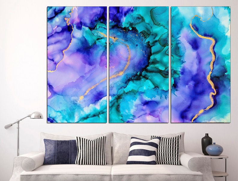 May include: Abstract wall art in a triptych arrangement. The artwork features a blend of blue, teal, and purple hues with gold accents. The fluid design creates a watercolor effect, adding a touch of elegance to any space. The artwork is displayed above a white sofa with decorative pillows.