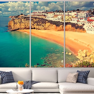 May include: A panoramic view of a coastal town in Portugal. The image shows white buildings perched on a cliff overlooking a sandy beach and turquoise water. The sky is blue with white clouds.
