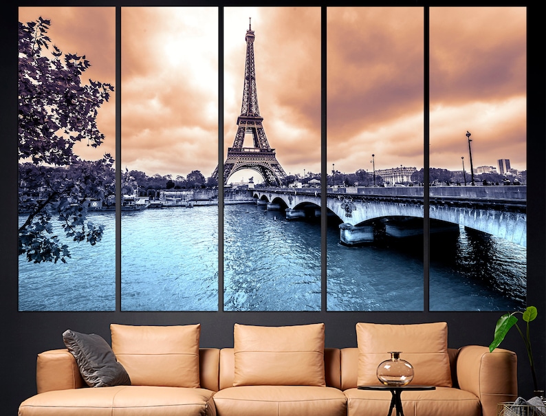 May include: A panoramic view of the Eiffel Tower in Paris, France. The tower is in the center of the image, with the Seine River and a bridge in the foreground. The sky is a soft orange and pink, with clouds.