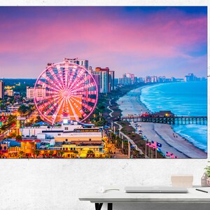 May include: A vibrant aerial view of Myrtle Beach, South Carolina at sunset. The iconic Ferris wheel stands out against the pink and purple sky, with the beach and ocean in the background. The pier extends into the water, and the city skyline is visible in the distance.