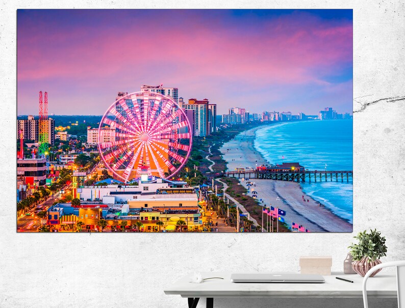 May include: A vibrant aerial view of Myrtle Beach, South Carolina at sunset. The iconic Ferris wheel stands out against the pink and purple sky, with the beach and ocean in the background. The pier extends into the water, and the city skyline is visible in the distance.