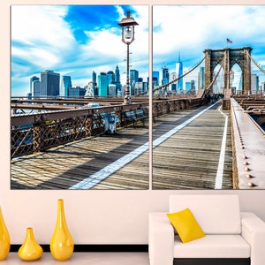 May include: A split canvas print featuring a view of the Brooklyn Bridge in New York City. The bridge is a suspension bridge with a brown wooden walkway and a view of the city skyline in the background. The sky is blue with white clouds.